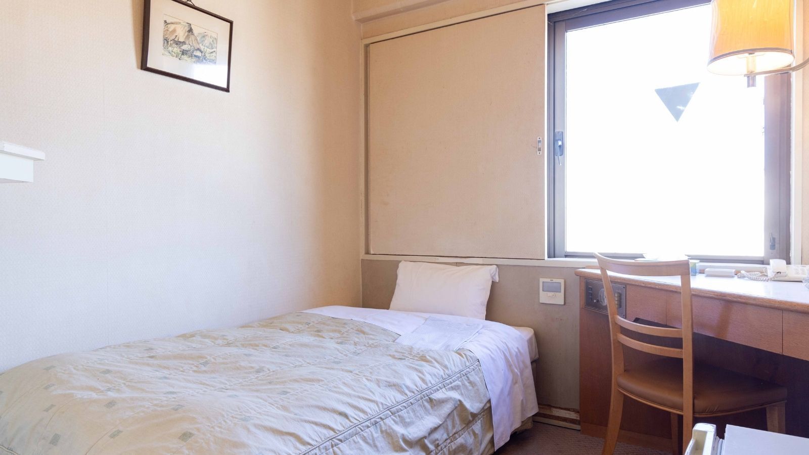 A single bed with a headboard, a desk with a chair, a window with a view, and a wall-mounted thermostat.