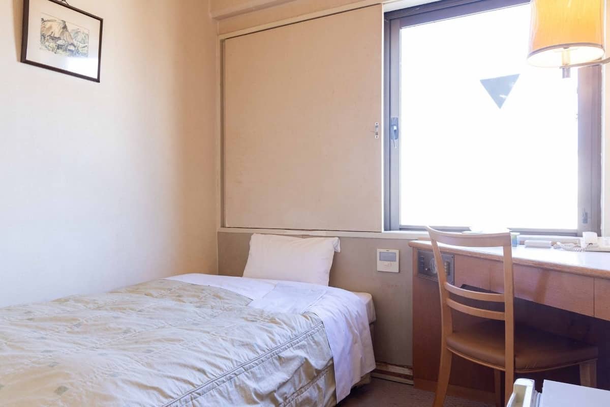 Uma cama de solteiro com cabeceira, uma escrivaninha com cadeira, uma janela com vista e um termostato fixado na parede.