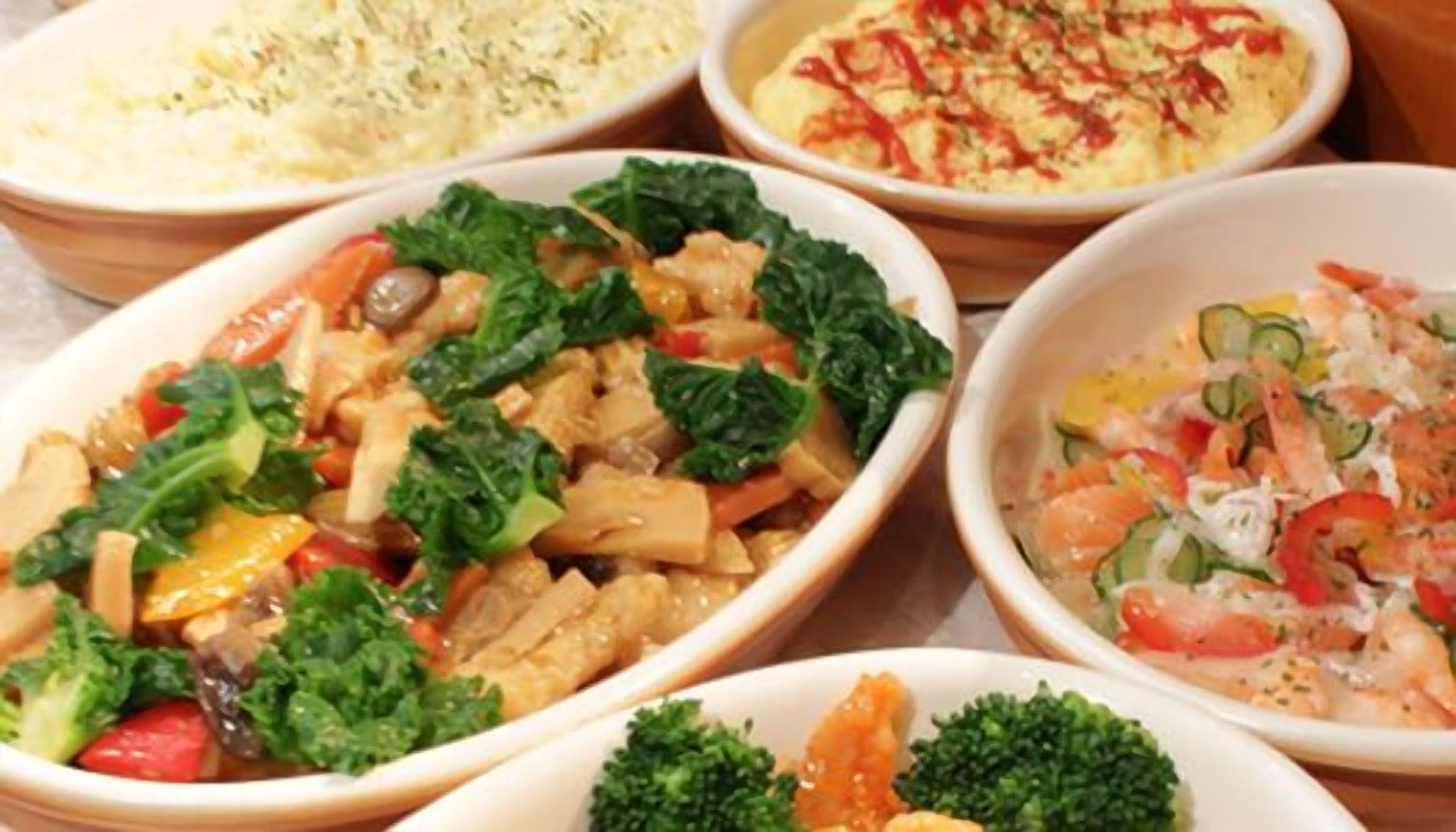 On trouve une variété de plats servis dans des bols, notamment une salade composée de légumes verts et de légumes, un plat de poulet et de légumes, et un plat agrémenté d’une sauce crémeuse.