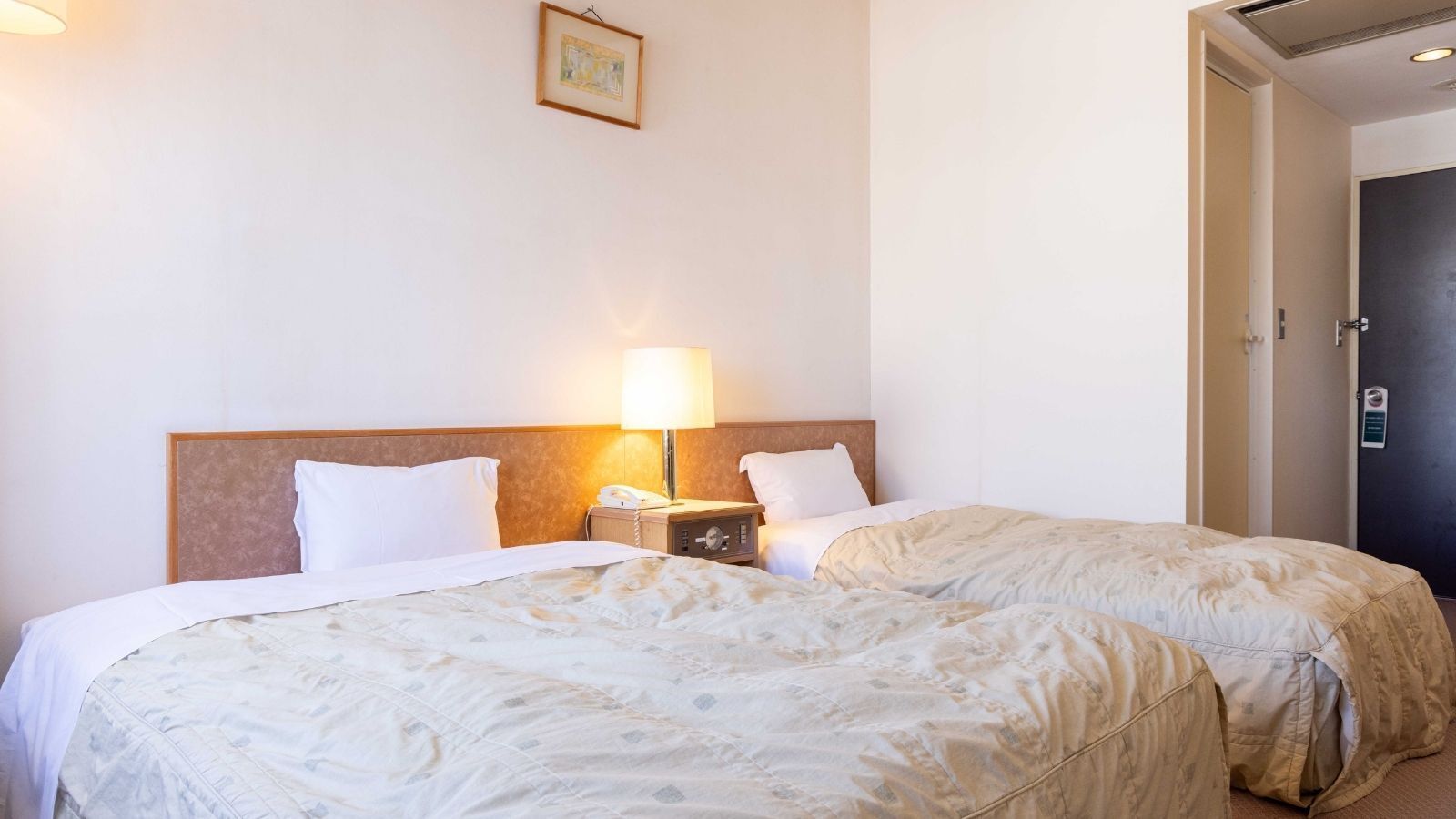 A hotel room with two beds, a nightstand with a lamp, and a door.