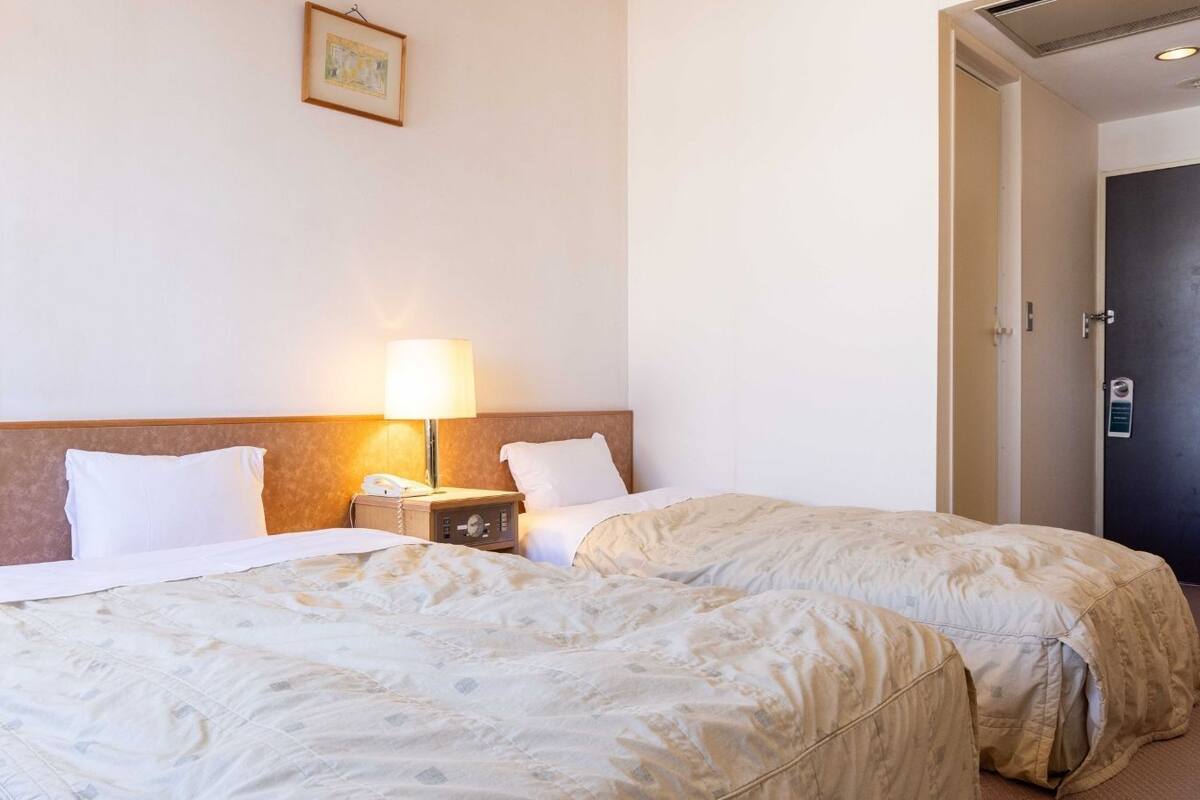 Quarto de hotel com duas camas, uma mesa de cabeceira com abajur e uma porta.