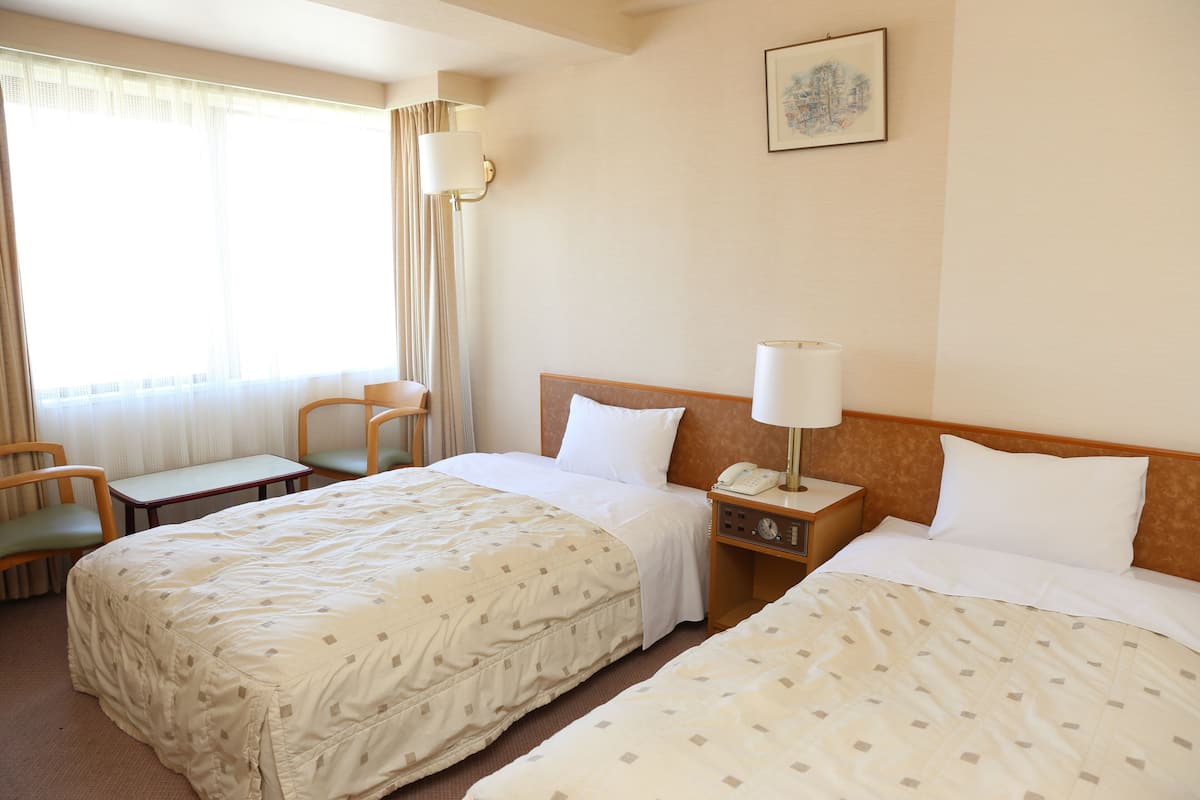 Quarto de hotel com duas camas, uma escrivaninha, uma cadeira, uma mesinha de cabeceira com abajur e um quadro na parede.