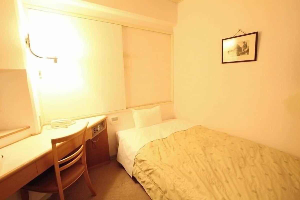 Quarto de hotel com cama, escrivaninha, cadeira, mesa de cabeceira e um quadro na parede.