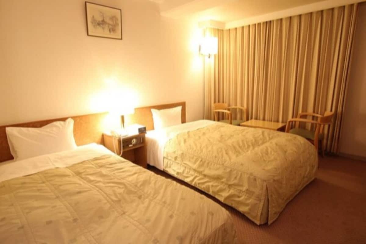 Quarto de hotel com duas camas, uma escrivaninha, uma cadeira, um abajur e uma cortina.