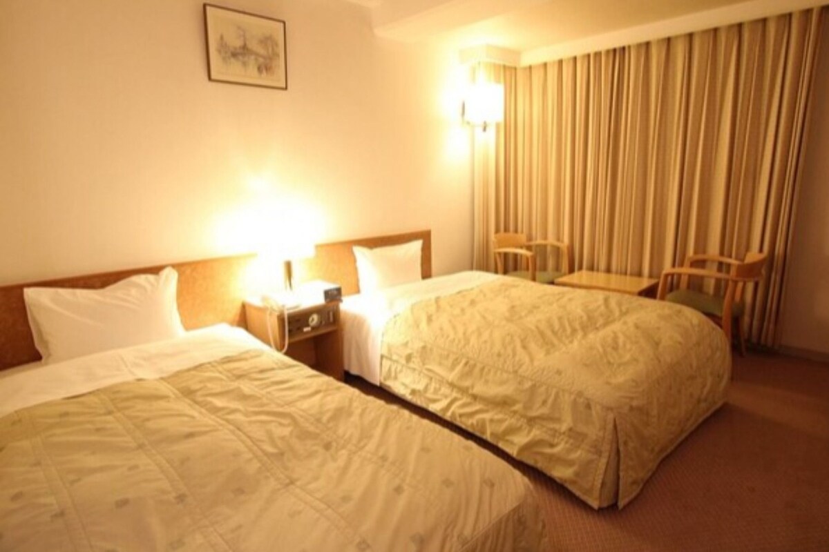 A hotel room with two beds, a desk, a chair, a lamp, and a curtain.
