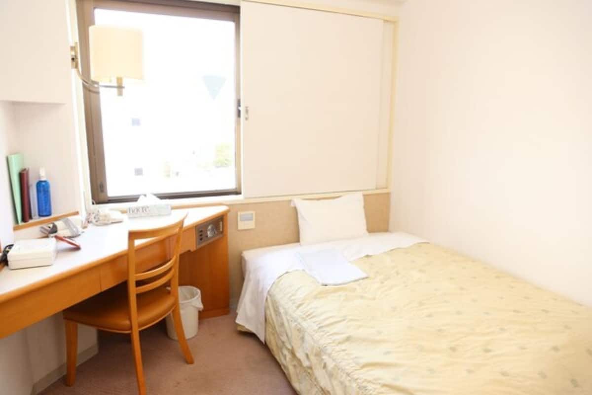 Quarto pequeno e arrumado com uma cama de solteiro, uma escrivaninha e uma cadeira. Há uma janela com vista para o exterior e um abajur sobre a mesa.