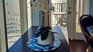 Une pièce avec vue sur la mer, une table ornée d’un vase de fleurs blanches et une chaise en bois.