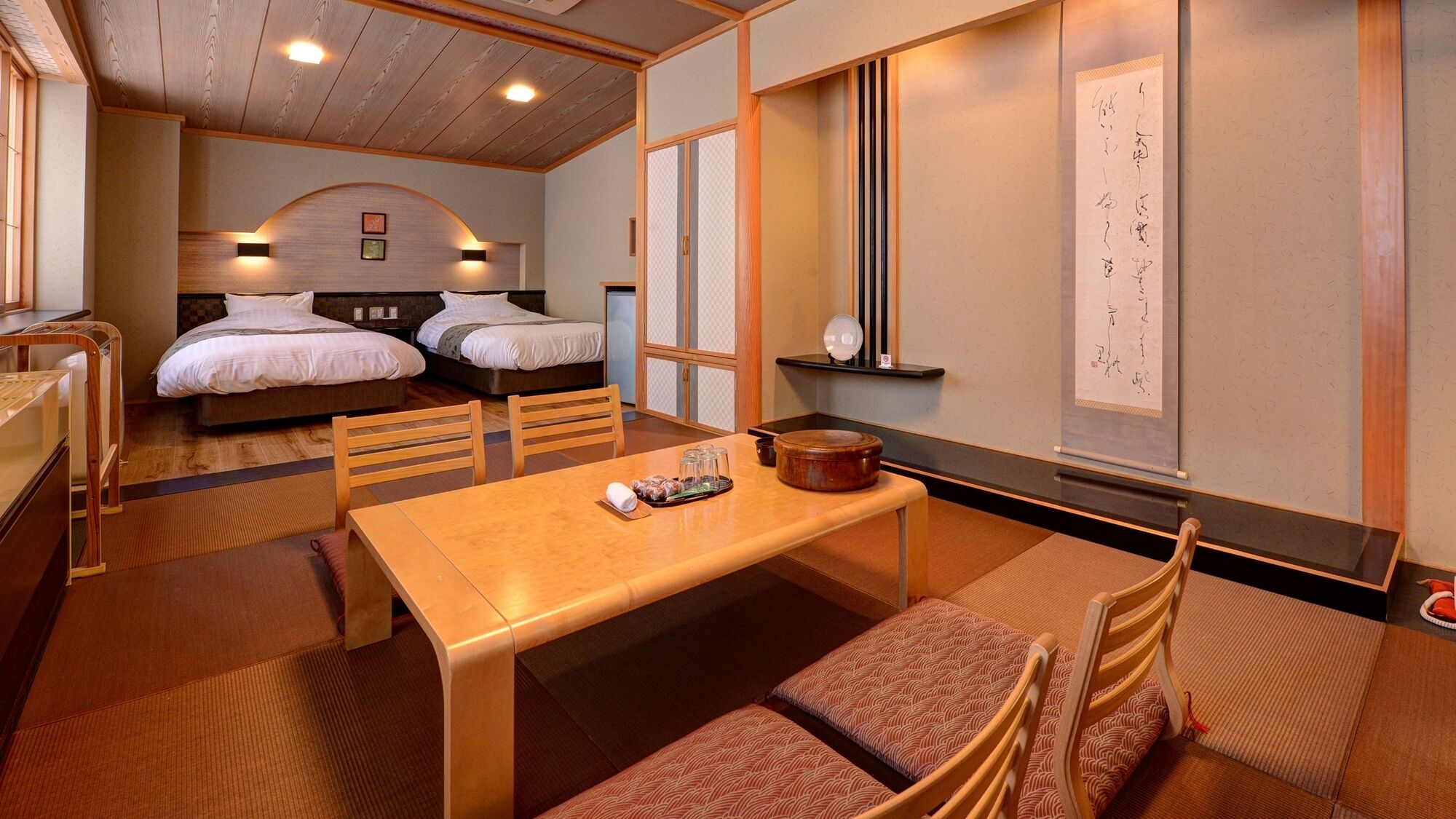 A hotel room with two beds, a wooden dining table, a small kitchen area, and a traditional Japanese writing on the wall.