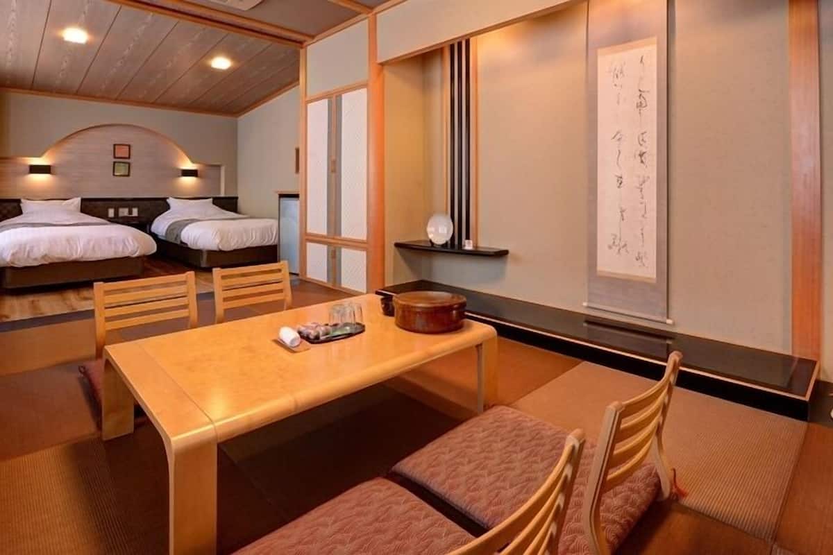 Egy szállodai szoba két ággyal, étkezőasztallal, székekkel, egy kis konyhasarokkal, és a falon található hagyományos japán kalligráfiai alkotással.