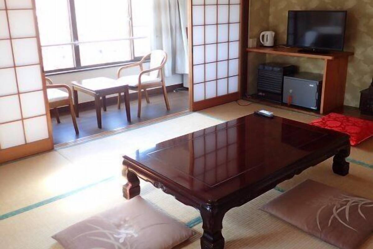 A traditional Japanese-style room with tatami flooring, a low wooden table, a television on a stand, and a sliding door leading to a balcony.