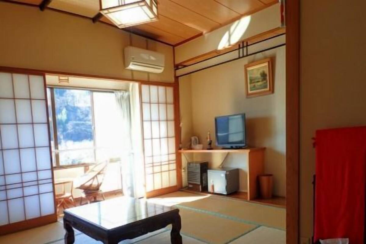 A traditional Japanese room with tatami flooring, a low table, a television, and a sliding door leading to an outdoor area.
