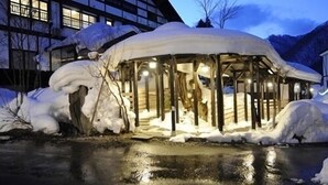 Une structure extérieure recouverte de neige, dotée d’un toit, peut-être un belvédère, située près d’un bâtiment avec de grandes fenêtres.