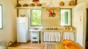 A compact kitchen with a white refrigerator, a small stove, and a window with a view of greenery. - Casa Jequitibá Rosa (Porto Seguro)
