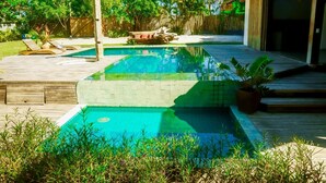 A rectangular swimming pool with a wooden deck surrounding it, a wooden pergola, and a fenced backyard with trees and plants. - Casa Jequitibá Rosa (Porto Seguro)