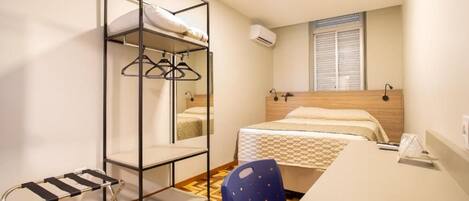 A room with a bunk bed, a desk with a chair, a hanging rack, and a window with blinds.