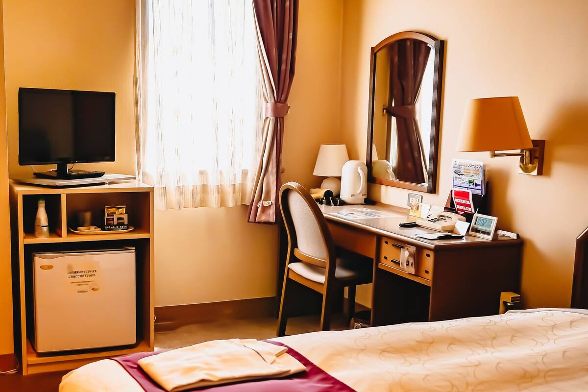 Una habitación de hotel con cama, escritorio con silla, televisión, refrigerador pequeño y ventana con cortinas.