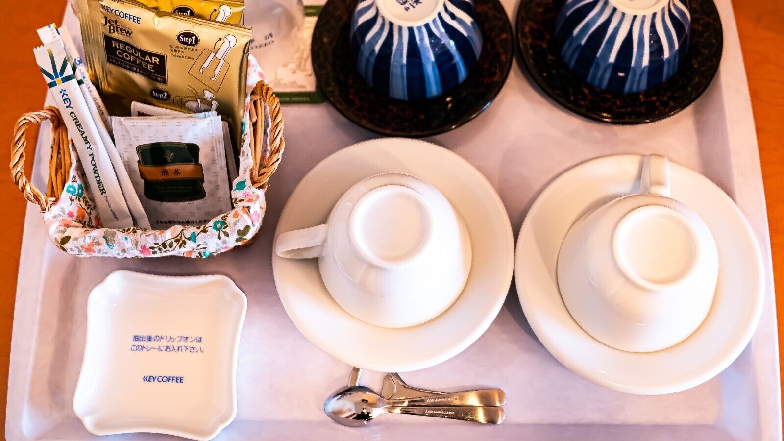 A tray with two white teacups, a spoon, a small dish with a label, and a basket containing coffee packets and a small container.