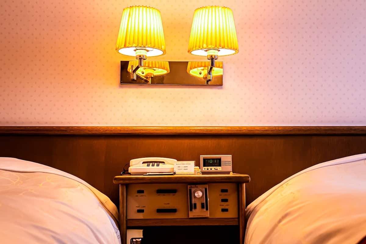 Habitación de hotel con dos camas, una mesita de noche con teléfono y reloj digital, y dos lámparas de pared.