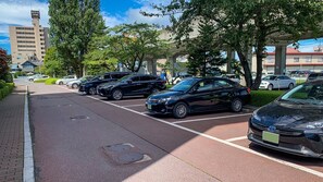 Un estacionamiento con varios coches, árboles y un edificio al fondo.