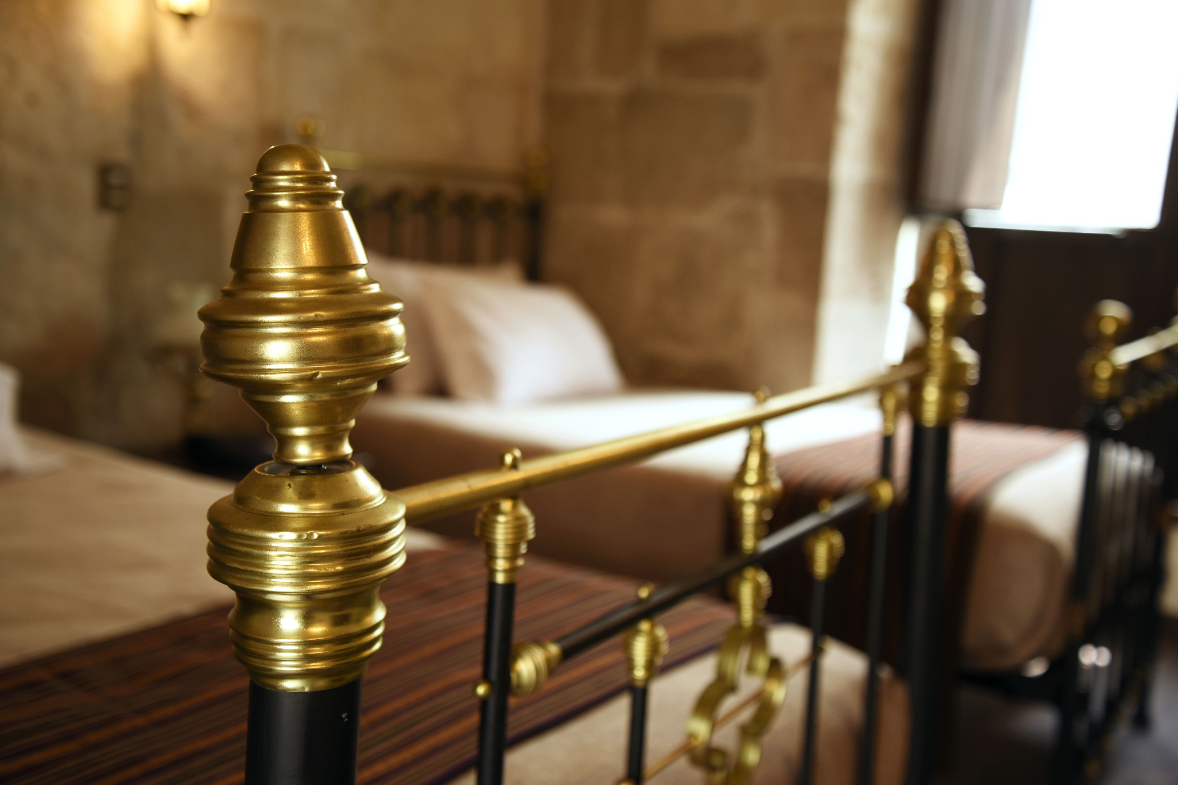 Una cama con estructura de madera y detalles en latón, una pared de piedra y una ventana con cortinas.