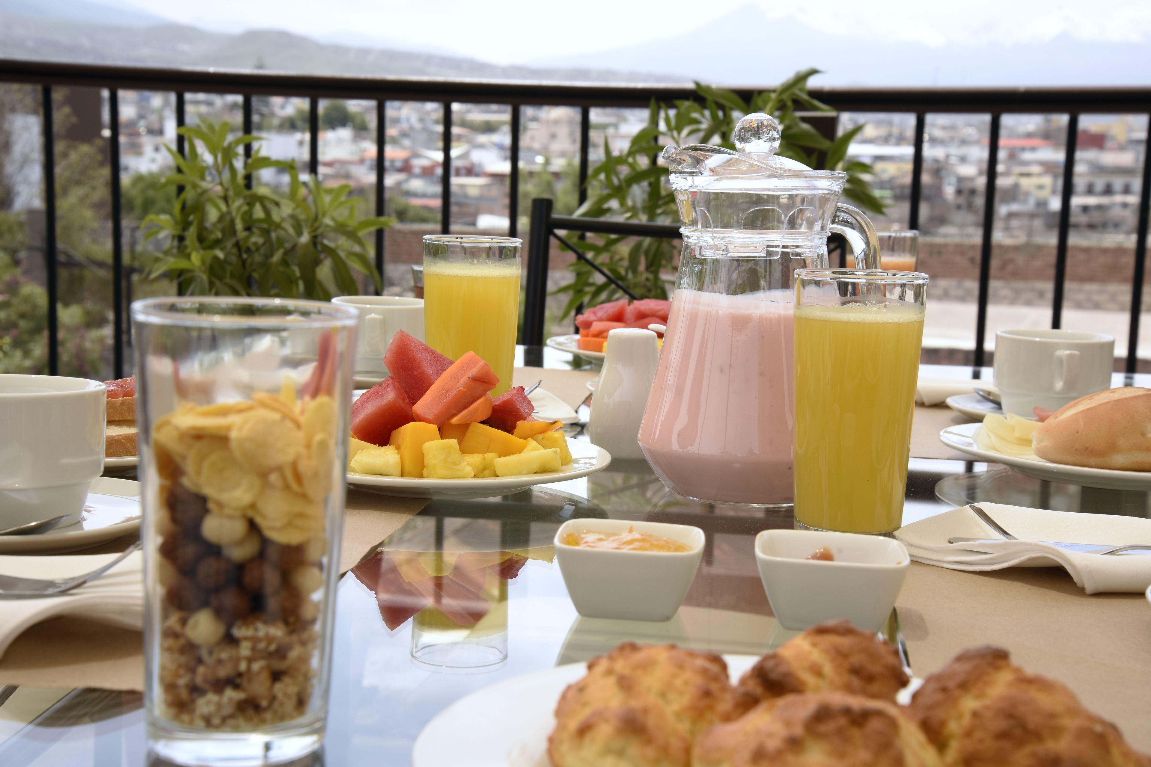 Une table de petit-déjeuner avec divers aliments et boissons, une carafe, et une vue sur la ville en arrière-plan.