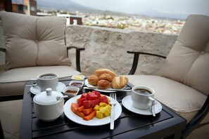 Una mesa con alimentos para desayuno, como café, pasteles y fruta, ubicada en un balcón con vista a la ciudad.