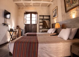 A room with two beds, a wooden headboard, a desk with a chair, a shelf with clothes, and a painting on the wall.