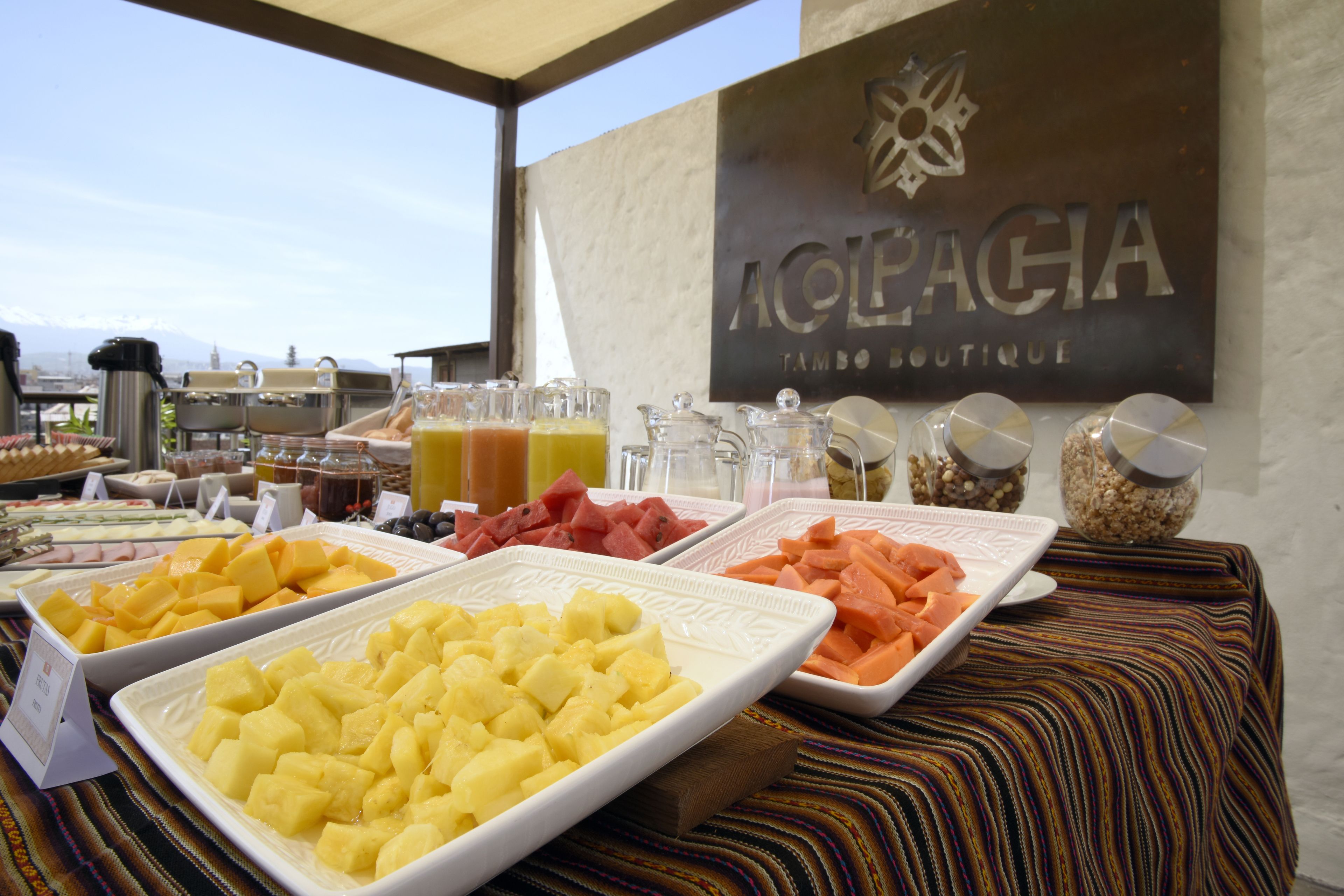 Une table de buffet garnie de divers fruits et boissons, une enseigne indiquant «Aolepaga Tango Boutique», et une vue sur des montagnes en arrière-plan.