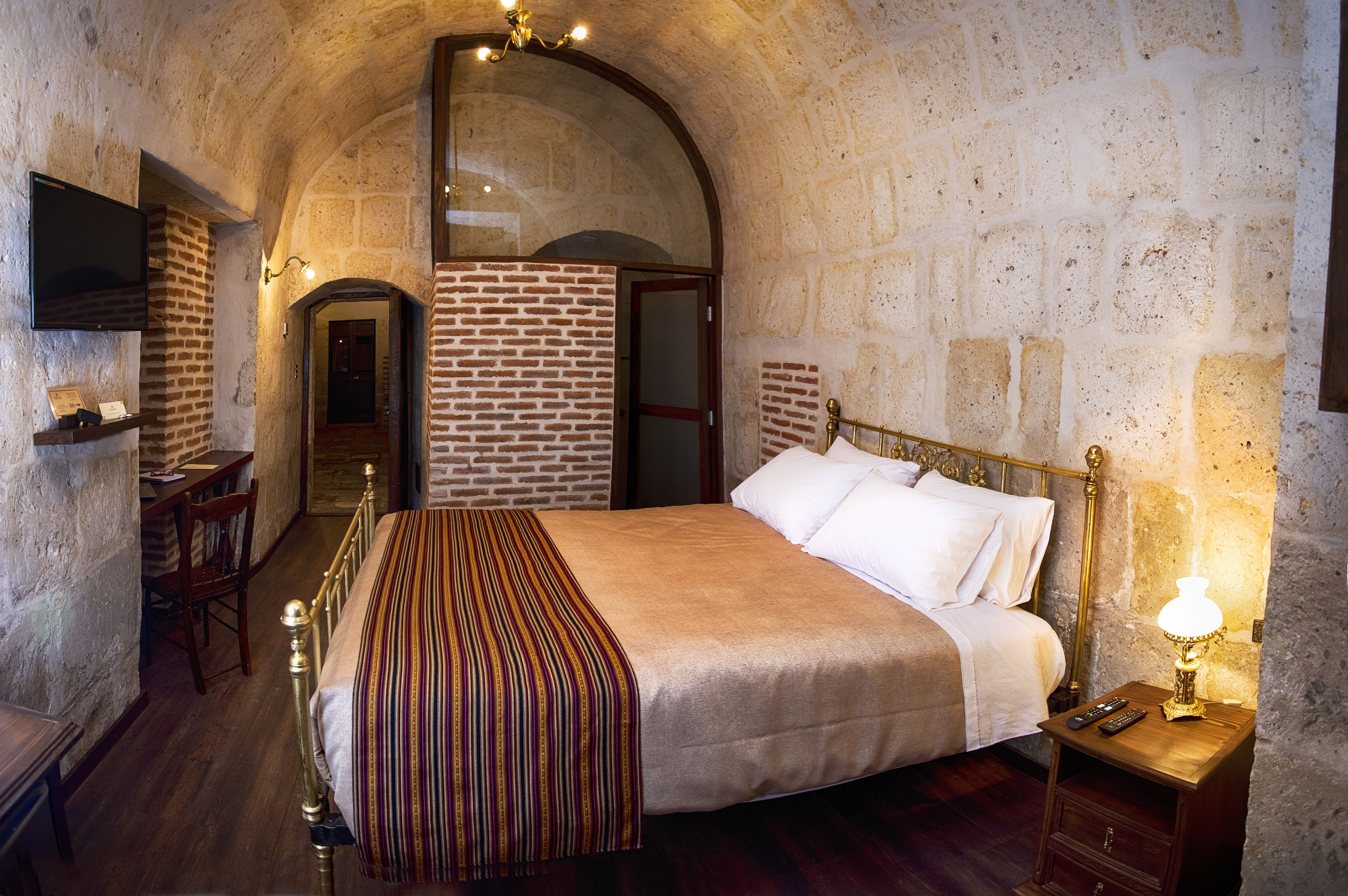 A bedroom with a brick wall, a wooden bed, a bedside table with a lamp, and a TV mounted on the wall.