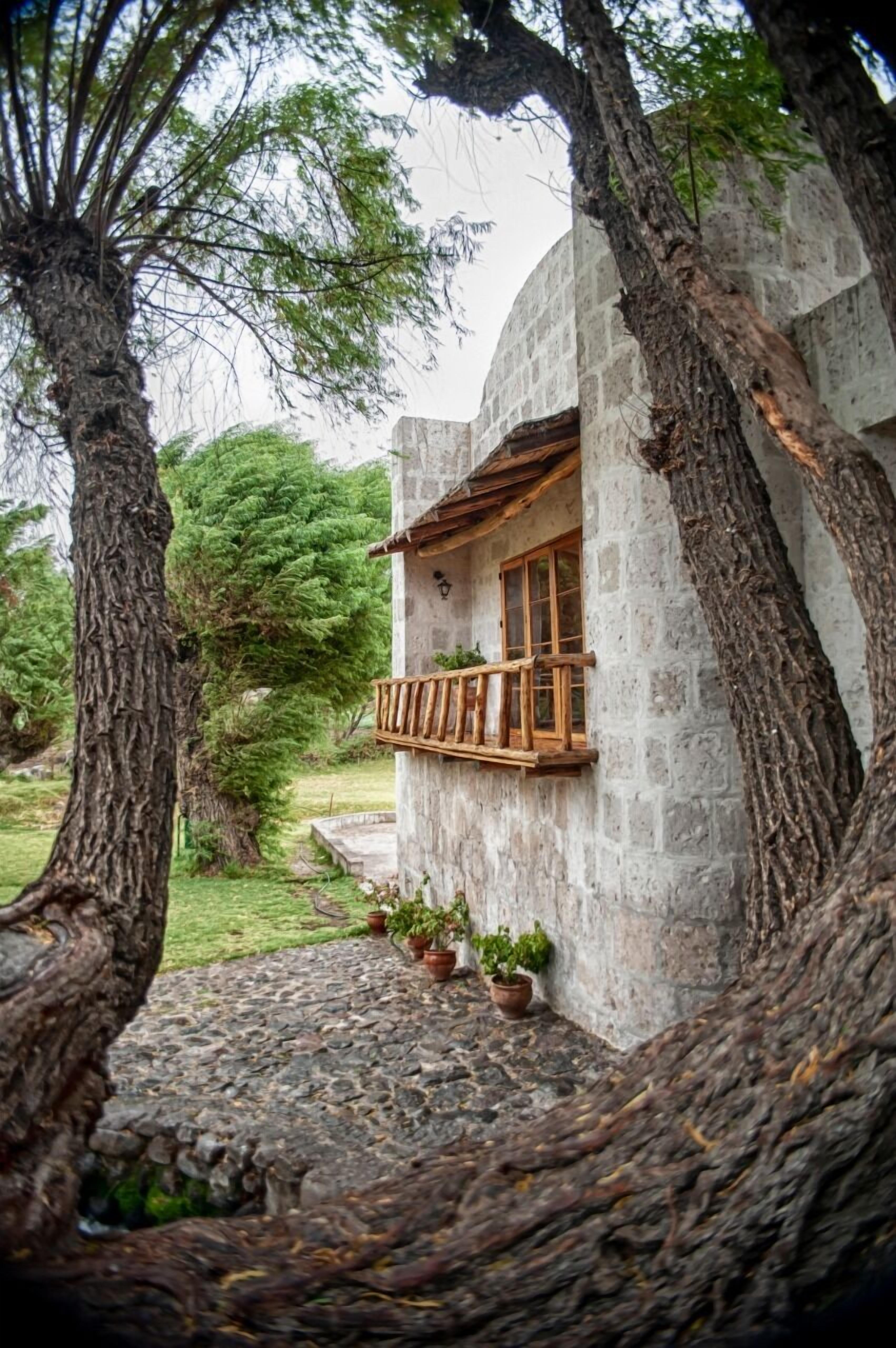 Edificio de piedra con balcón de madera, rodeado de árboles y plantas en macetas.