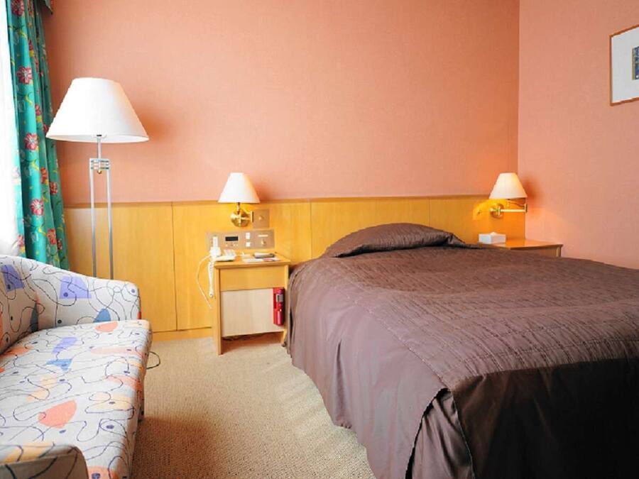 Une chambre d’hôtel avec un lit, une table de chevet avec une lampe, un petit bureau avec un ordinateur et un canapé à motifs.