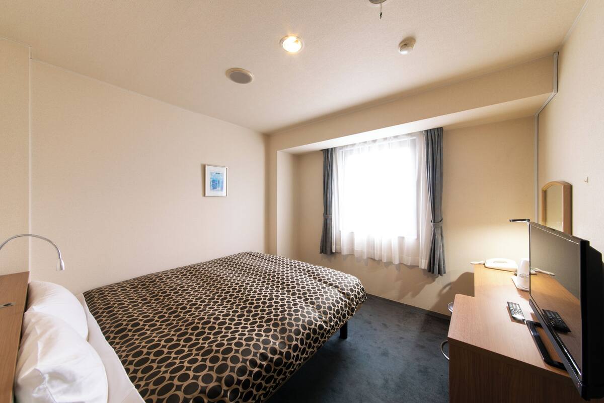 A hotel room with a bed, a desk with a computer, a mirror, and a window with curtains.