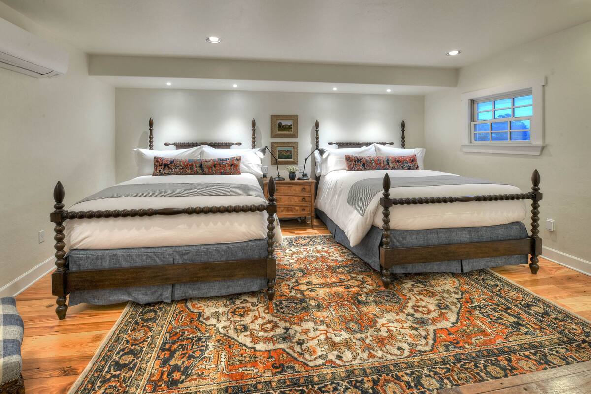 A bedroom with two beds, a large patterned rug, wooden floors, and a window with a view.