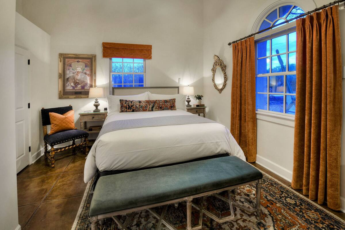 A bedroom with a large bed, a bench, a nightstand, a lamp, a framed picture, and a window with curtains.