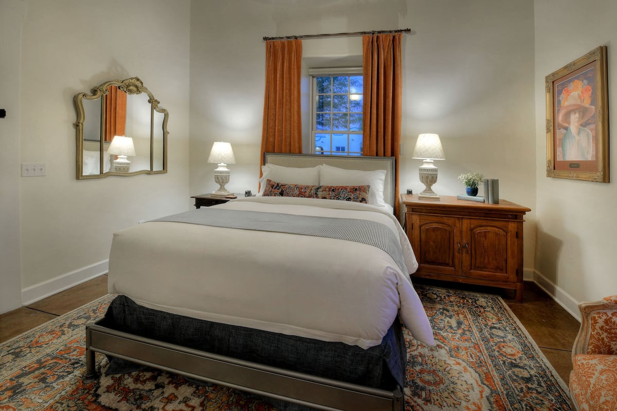 A neatly arranged bedroom with a bed, two bedside tables with lamps, a nightstand, a wooden cabinet, and a large mirror on the wall.
