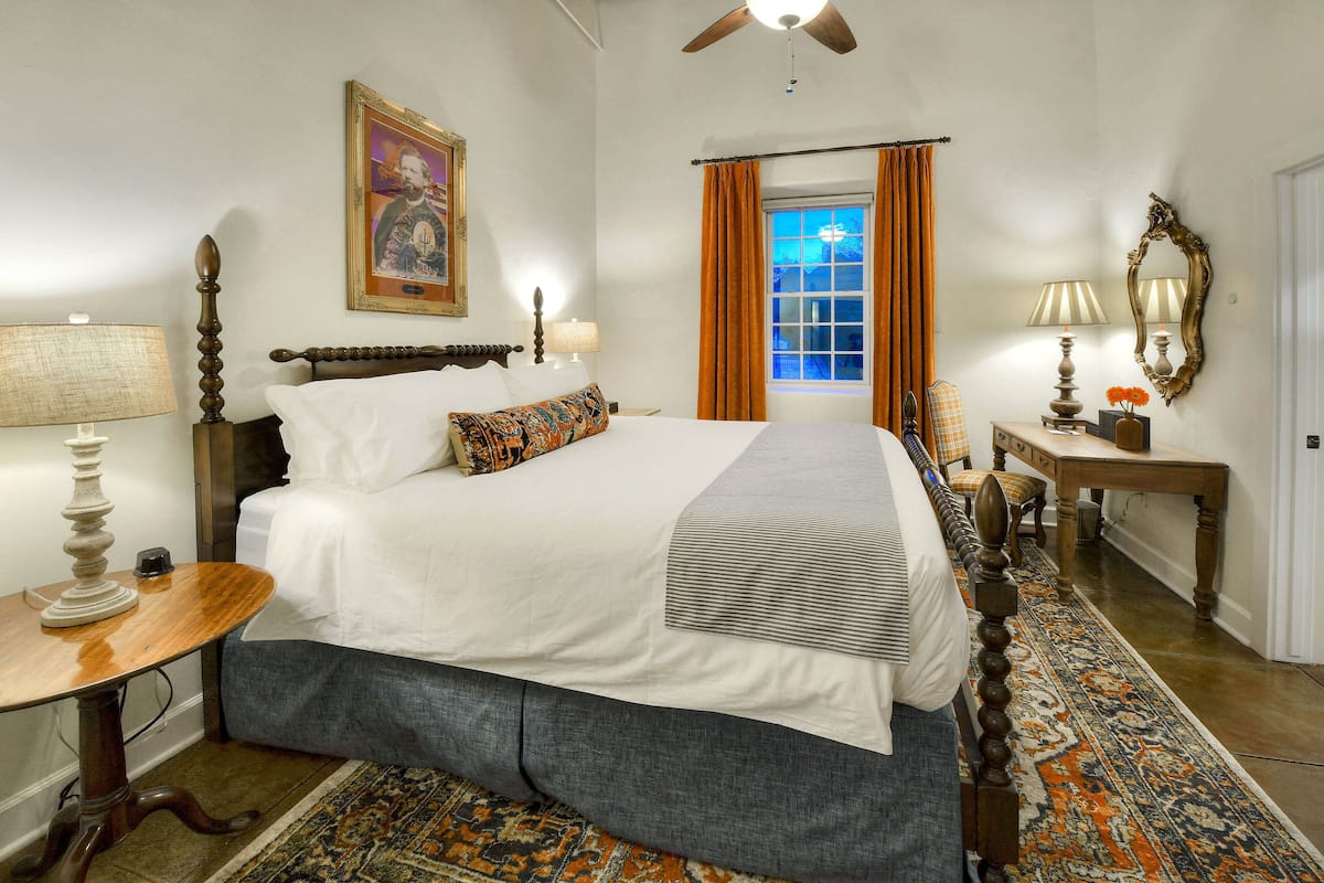 A hotel room with a large bed, a wooden nightstand, a table lamp, a mirror, and a window with curtains.