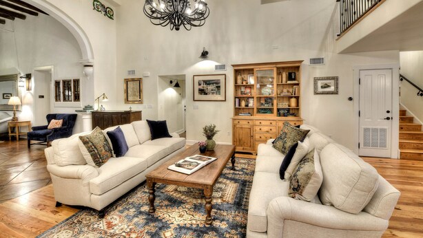 A spacious living room with two sofas, a coffee table, and a patterned rug. There is a chandelier, a wooden cabinet with decorative items, and a staircase in the background.