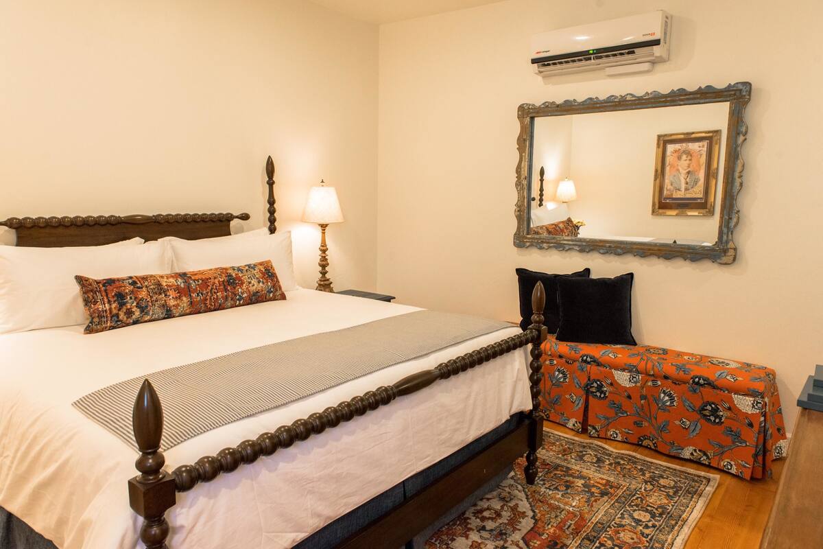 A neatly made bed with a wooden headboard, a bedside table with a lamp, a small bench with a floral bedspread, and a large mirror with a picture frame.