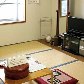 Une pièce avec un sol recouvert de tatamis, une télévision, un radiateur et une petite table sur laquelle se trouvent des papiers.