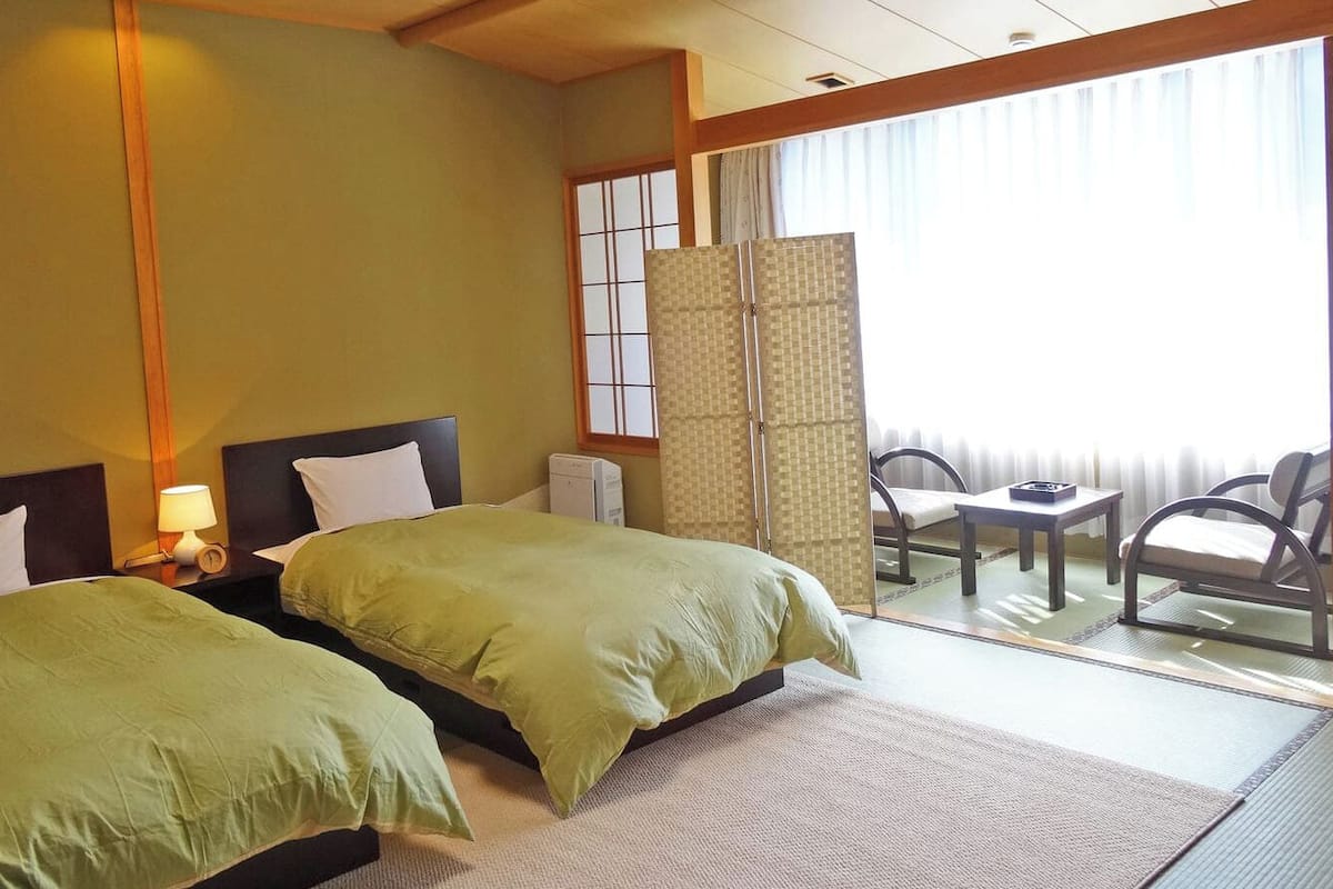 A hotel room with two beds, a small table, a chair, and a view through a sliding door.