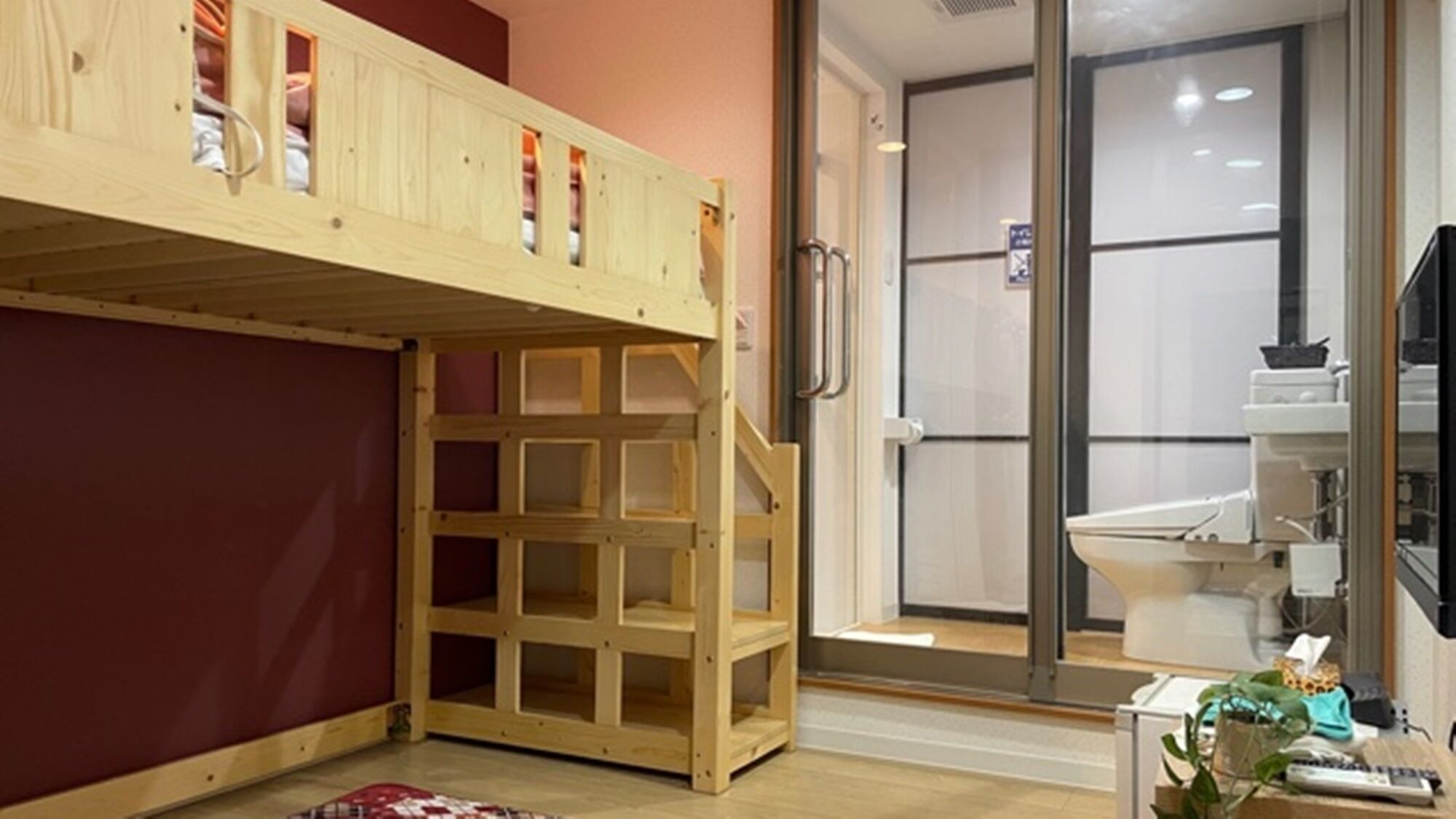 Una habitación con una cama litera, un vidrio que da a un baño y una mesita con una planta.