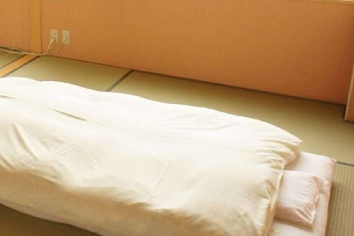 A single bed with a white mattress and pink pillow in a room with a tiled floor and peach-colored walls.