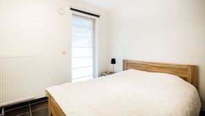 A bedroom with a bed, a radiator, a window with blinds, and a nightstand with a lamp. - Santos Secundo GV01 (Koksijde)