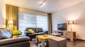 A modern living room with a leather sofa, a wooden coffee table, a TV on a wooden cabinet, and a large window with curtains. - Santos Secundo GV01 (Koksijde)