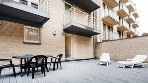 A rooftop patio with a table and chairs, a lounge area with sun loungers, and a brick building with balconies. - Santos Secundo GV01 (Koksijde)