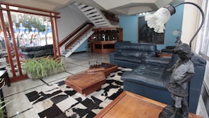 Un salón amplio con un sofá de cuero negro, una mesa de centro de madera y una estatua. Se ve una escalera al fondo, que conduce a un piso superior.