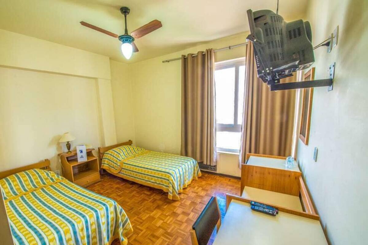 Quarto com duas camas, ventoinha no teto, televisão na parede e janela com cortinas.