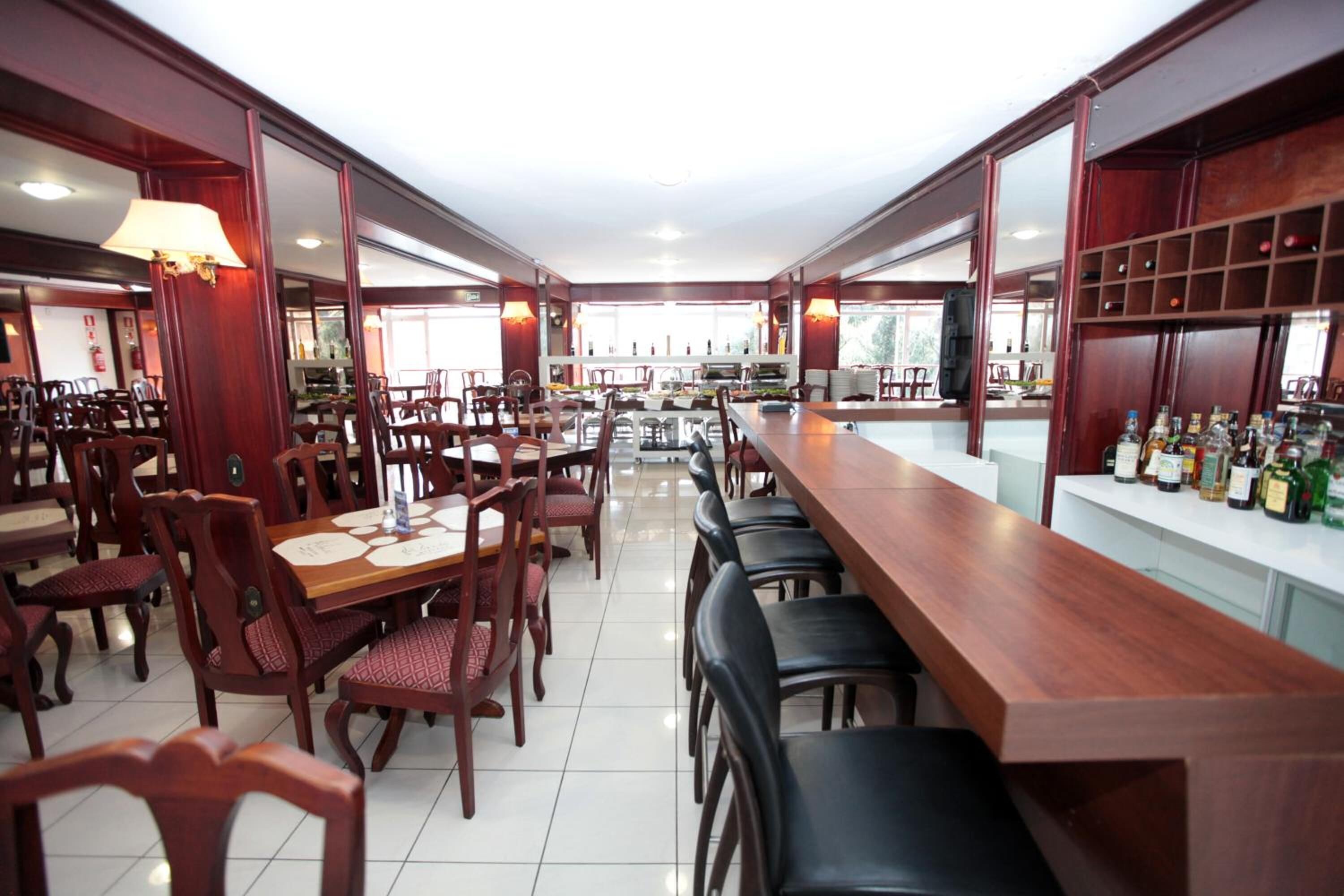 Um restaurante espaçoso com várias mesas e cadeiras, um balcão comprido com área de bar e vista para o exterior através de grandes janelas.