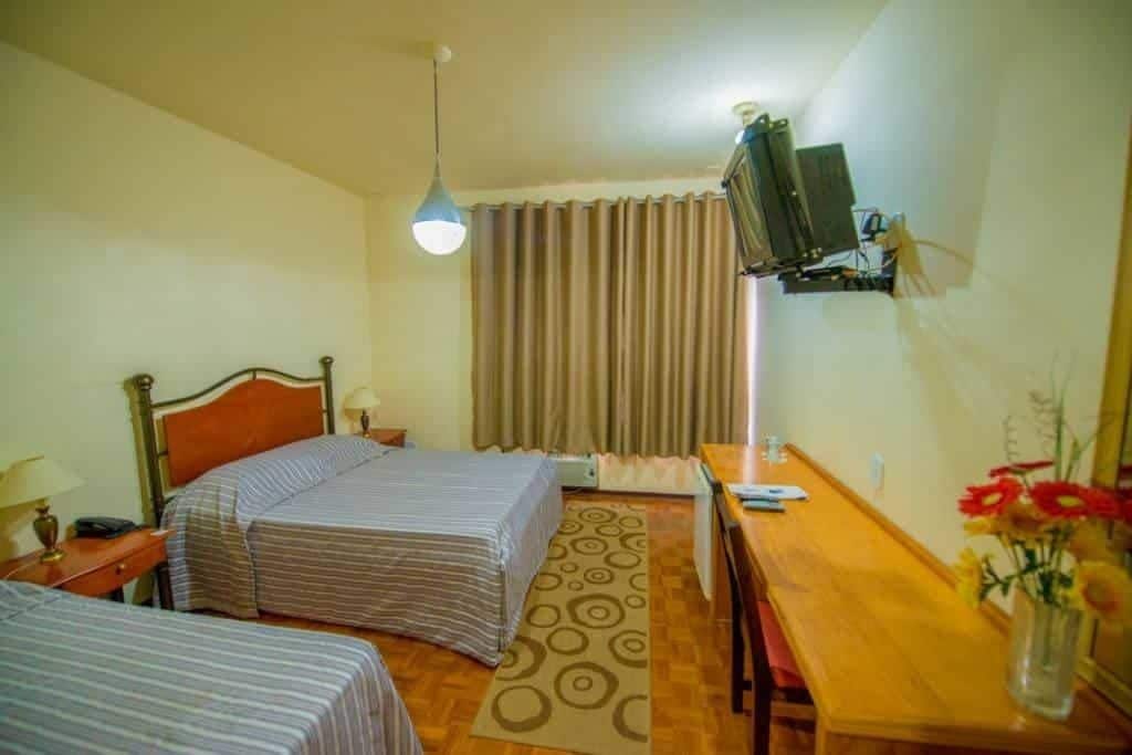 A hotel room with two beds, a wooden desk, a television mounted on the wall, and a window with curtains.