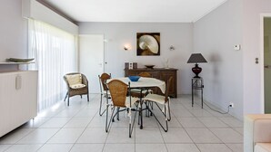 A dining area with a white table, wicker chairs, a blue bowl, a lamp, and a painting on the wall. - Etoilée GV04 (Koksijde)